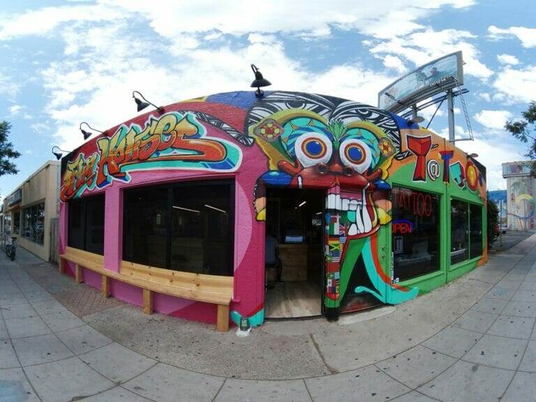 A brightly colored building with vibrant murals, featuring a cartoonish face, "Tattoos" sign, and graffiti art. The street scene includes paved sidewalks and a fish-eye lens effect.