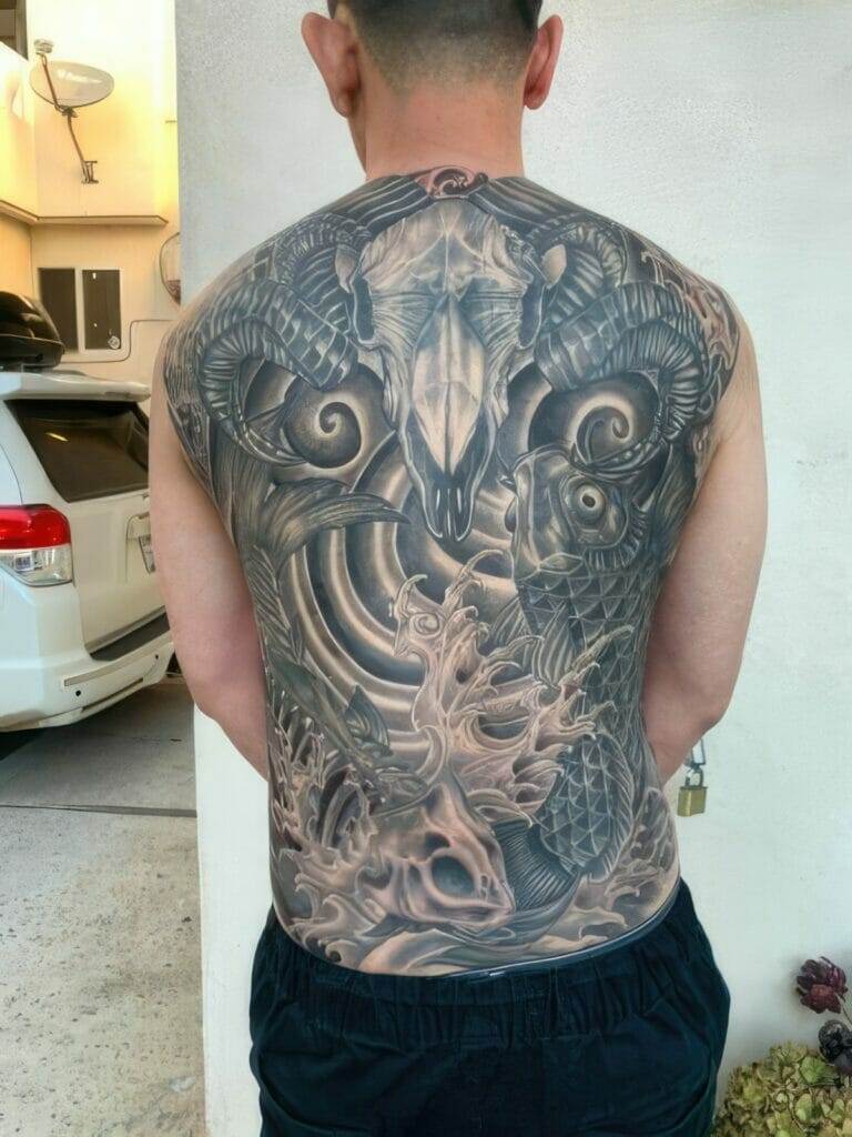 A person with a large, intricate tattoo covering their entire back, featuring a ram skull, fish scales, and various detailed elements. The individual is standing outside next to a white vehicle.