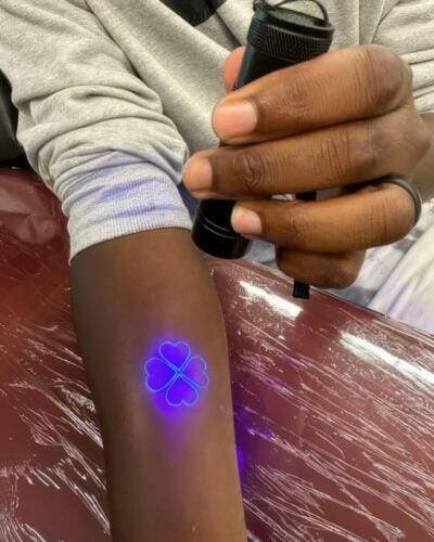 A person holds a UV flashlight over their arm, revealing a glowing blue four-leaf clover design.