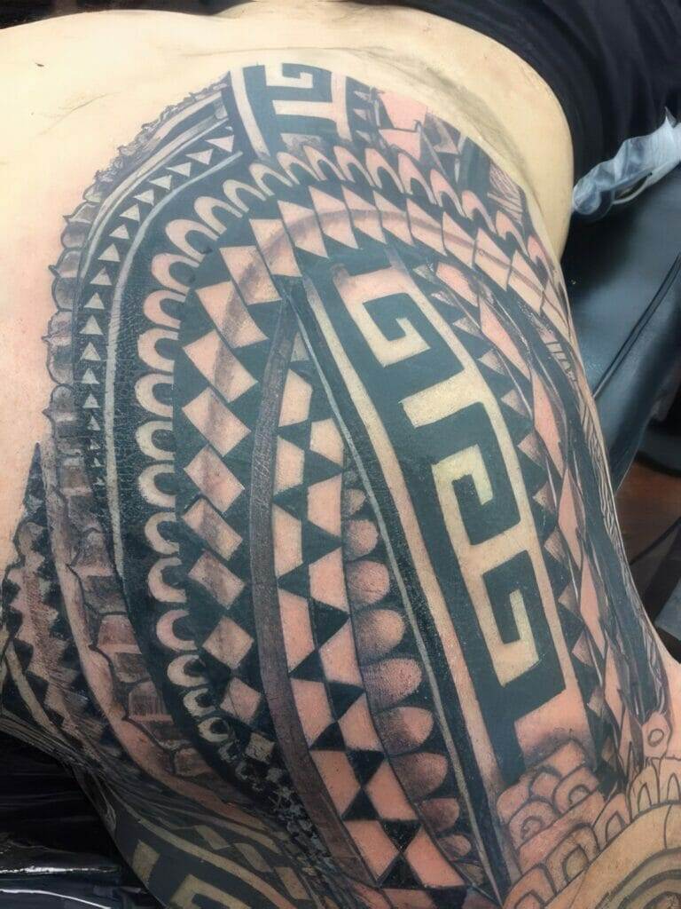 A close-up of an intricate black and grey tribal tattoo covering a person's side and part of their back. The design includes geometric patterns and symbols.