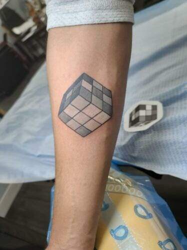 A forearm with a tattoo of a black, white, and gray Rubik's cube.