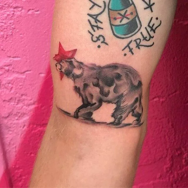 A tattoo of a bear on an arm, with the bear wearing a red star on its head. Other tattoos are visible in the background.