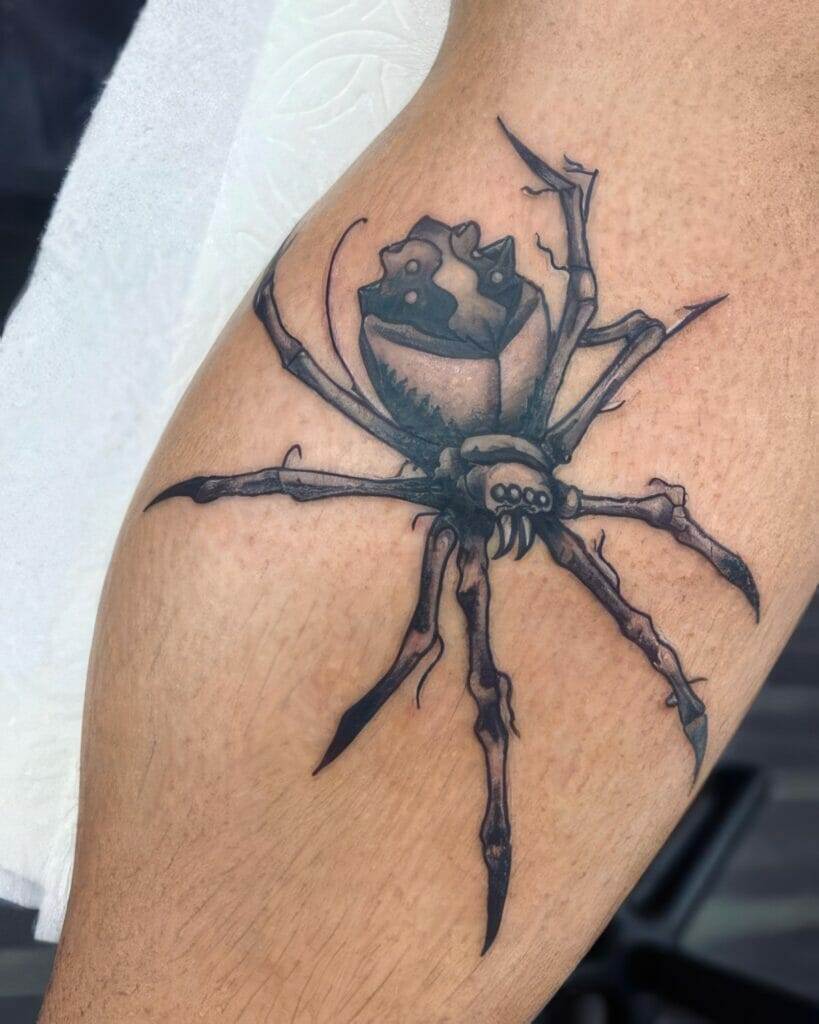 A tattoo of a detailed spider is inked on a person's skin. The spider is depicted with elongated legs and a detailed body on a light skin tone.