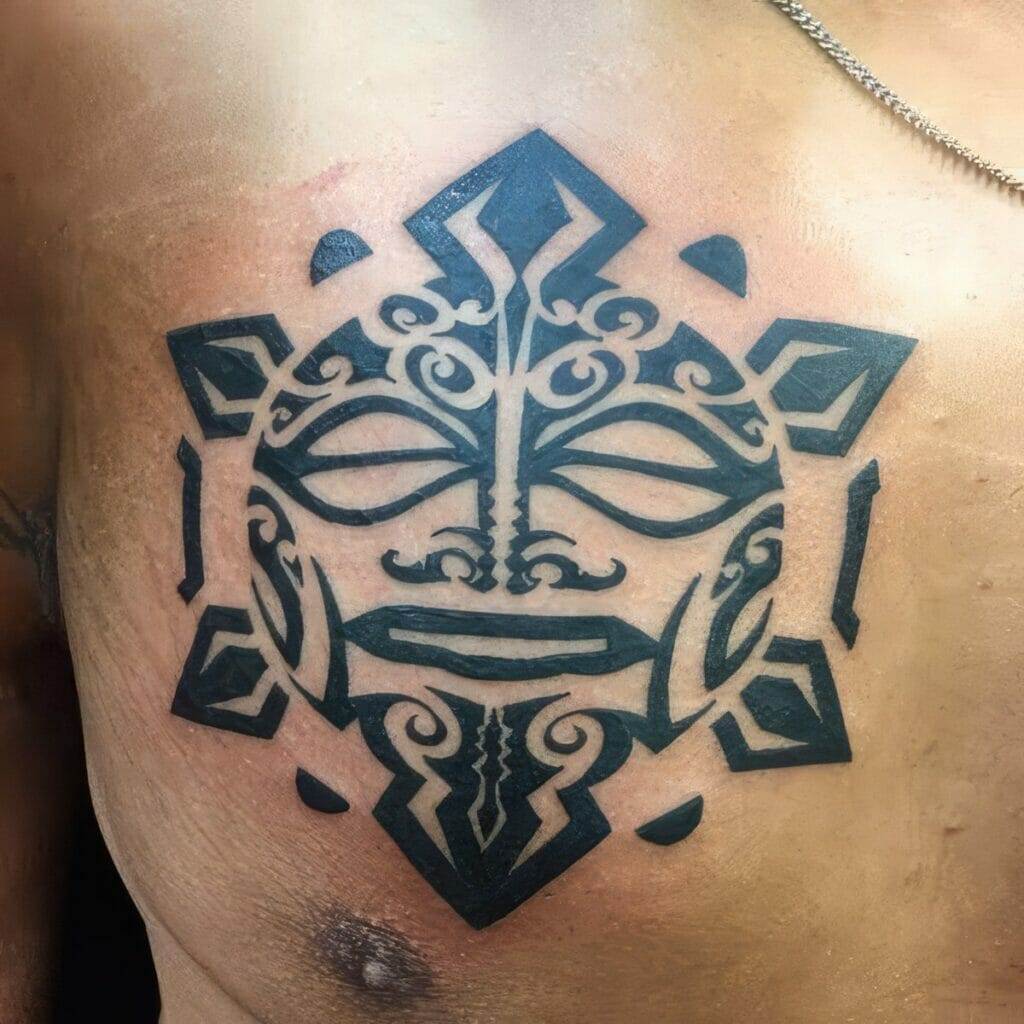 A black tribal mask tattoo on the upper chest of a person. The design features symmetrical patterns and geometric shapes.