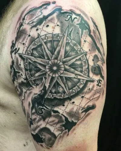 A detailed, grayscale tattoo of a compass rose with directional markers (N, S, E, W) is inked on a person's upper arm, featuring an artistic map-like background.