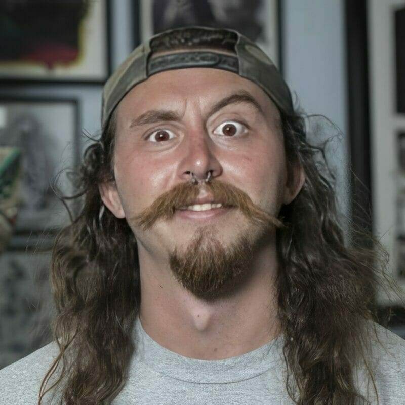 Man with long curly hair, mustache, beard, wearing a backwards cap, smiling.