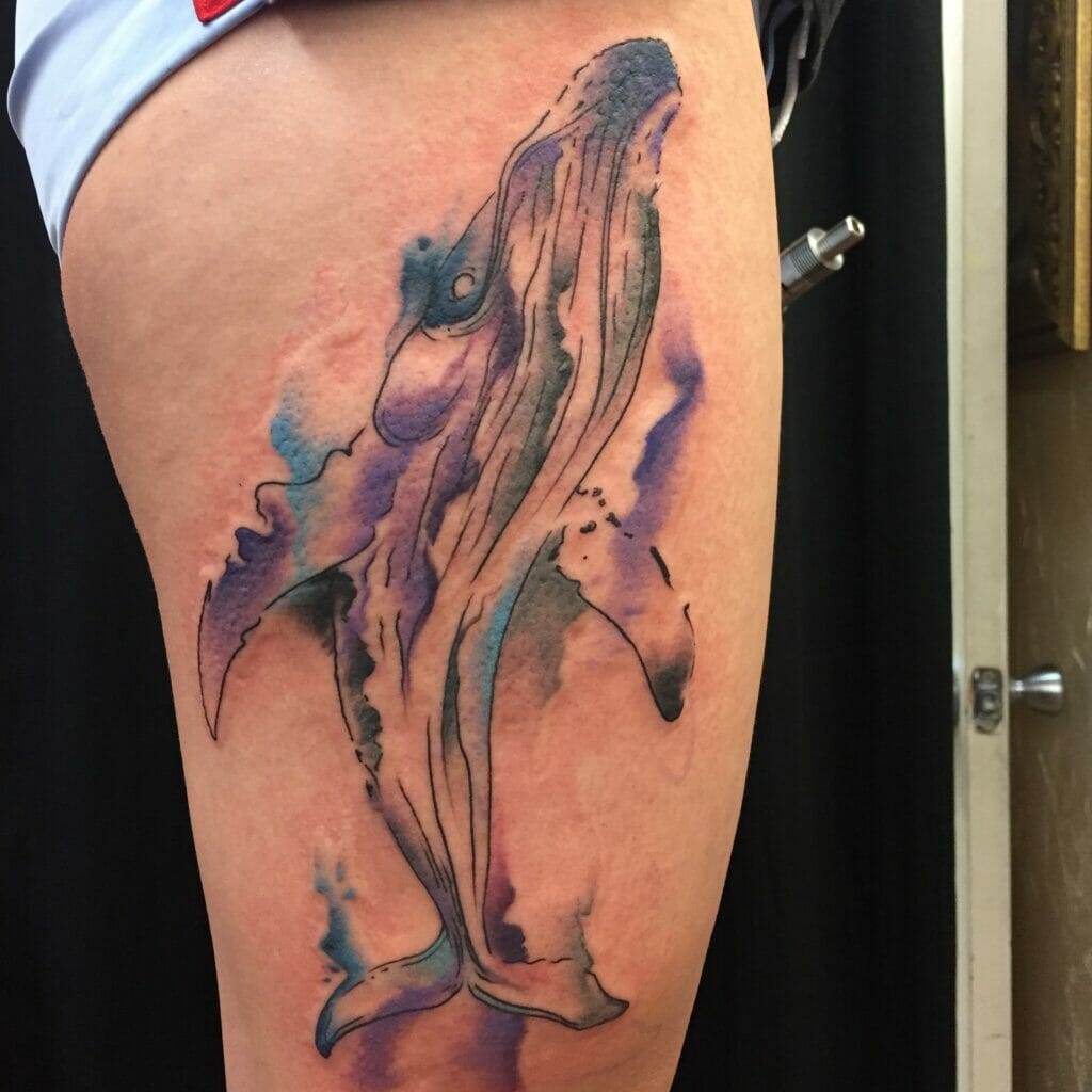 Tattoo of a whale with watercolor-style splashes in blue and purple on the upper thigh.