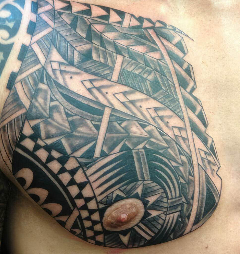 A close-up of a person's chest showing a large, detailed black-and-gray geometric tattoo surrounding the nipple.