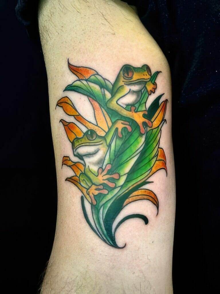 A colorful tattoo of two green and yellow frogs sitting on orange and green leaves, located on an upper arm.