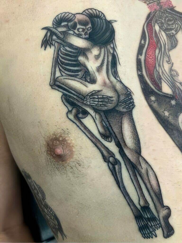 A tattoo depicting a skeleton embracing a nude woman with horns on her head, positioned on the skin near a human nipple.