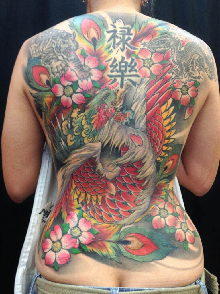 A person displays a large, colorful back tattoo featuring a phoenix, flowers, Chinese characters, and a tiger.