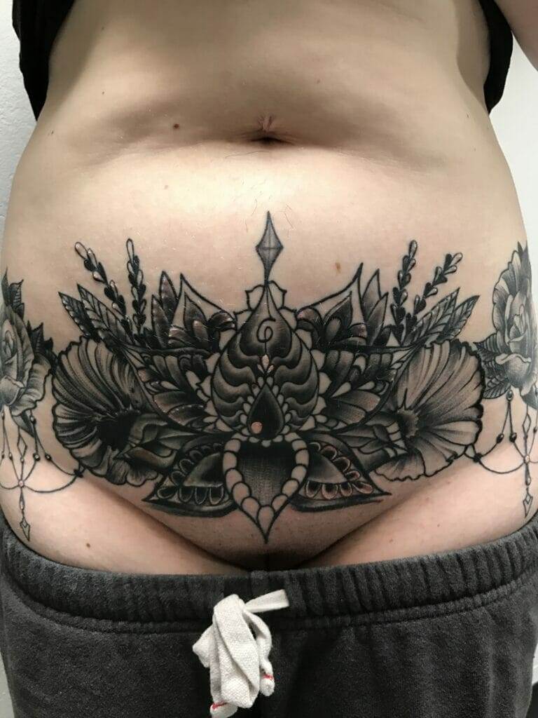 A detailed black ink tattoo featuring a symmetrical floral design is displayed on a person's lower abdomen.