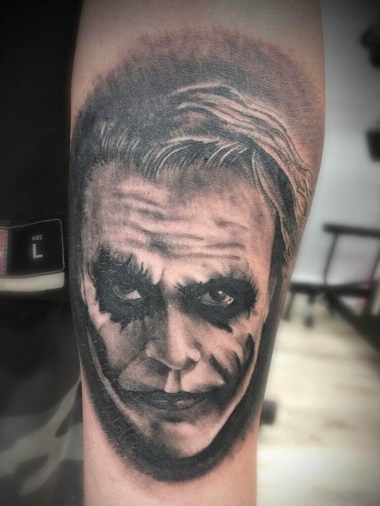 Forearm tattoo depicting a monochrome portrait of a character with dark eye makeup and a sinister smile.