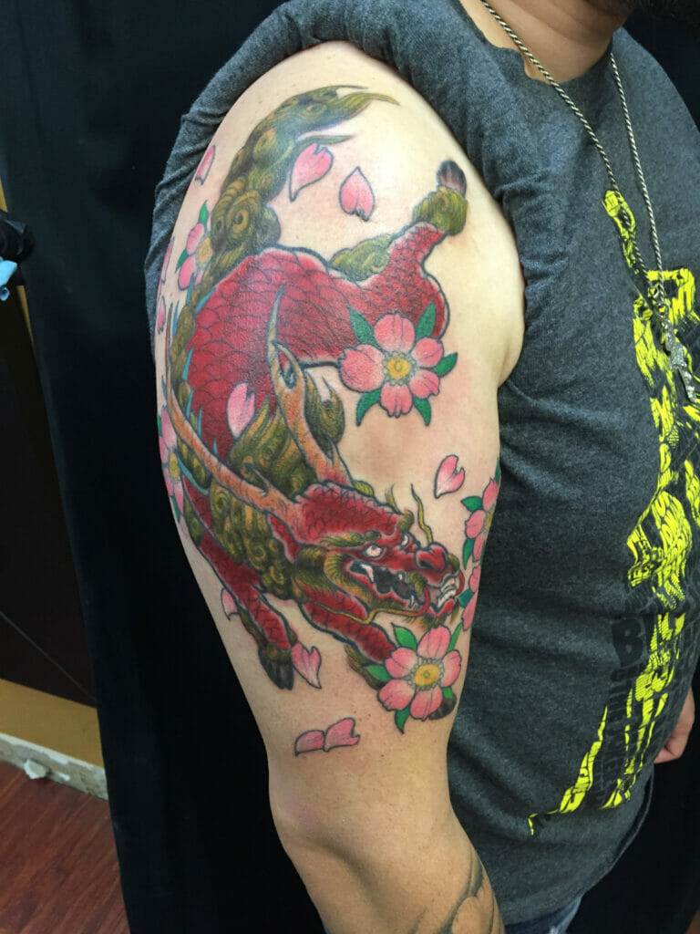 A person displays a detailed upper arm tattoo featuring a red dragon intertwined with flowers and petals.