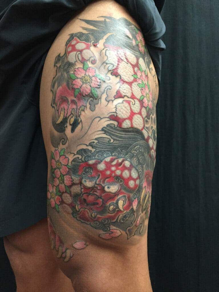 A person displays a detailed leg tattoo featuring a dragon with red scales, surrounded by pink flowers and black swirling patterns.