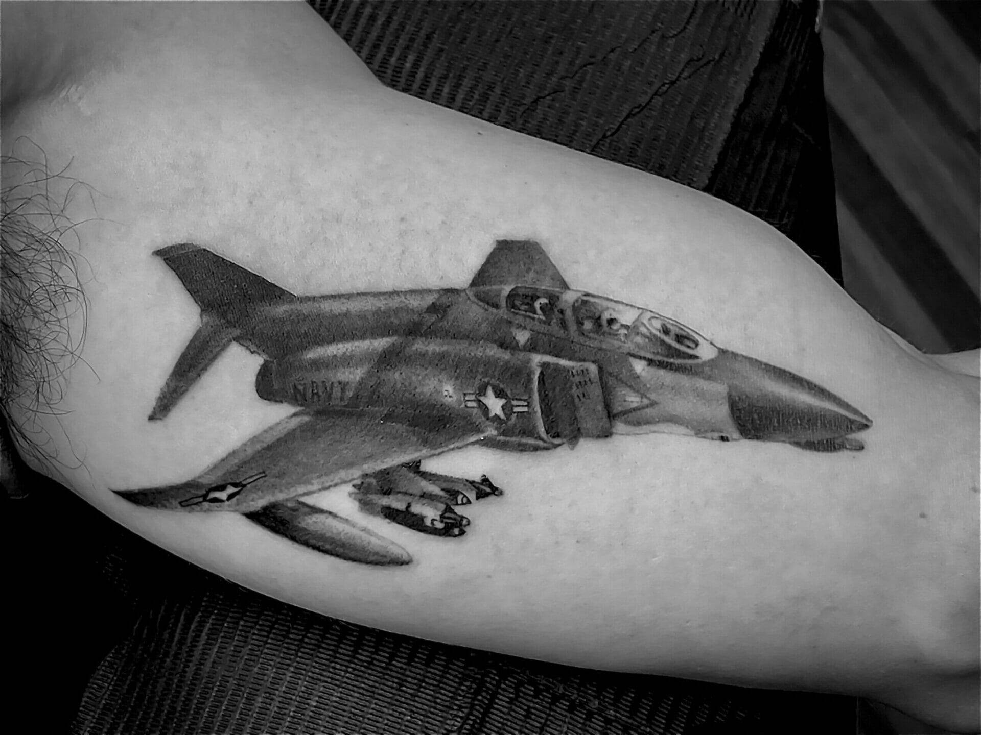 A black and white tattoo of a military jet with missiles, marked with "Navy" and other symbols, on a person's upper arm.