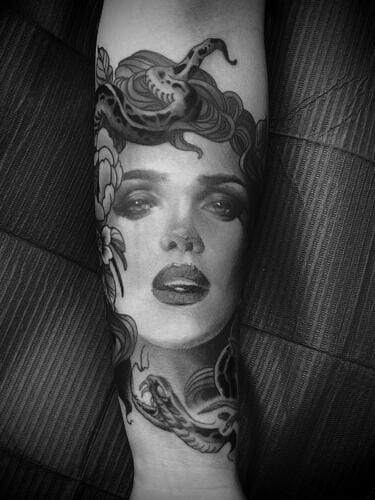 A black and gray tattoo on an arm depicting a woman's face surrounded by snakes and floral designs.