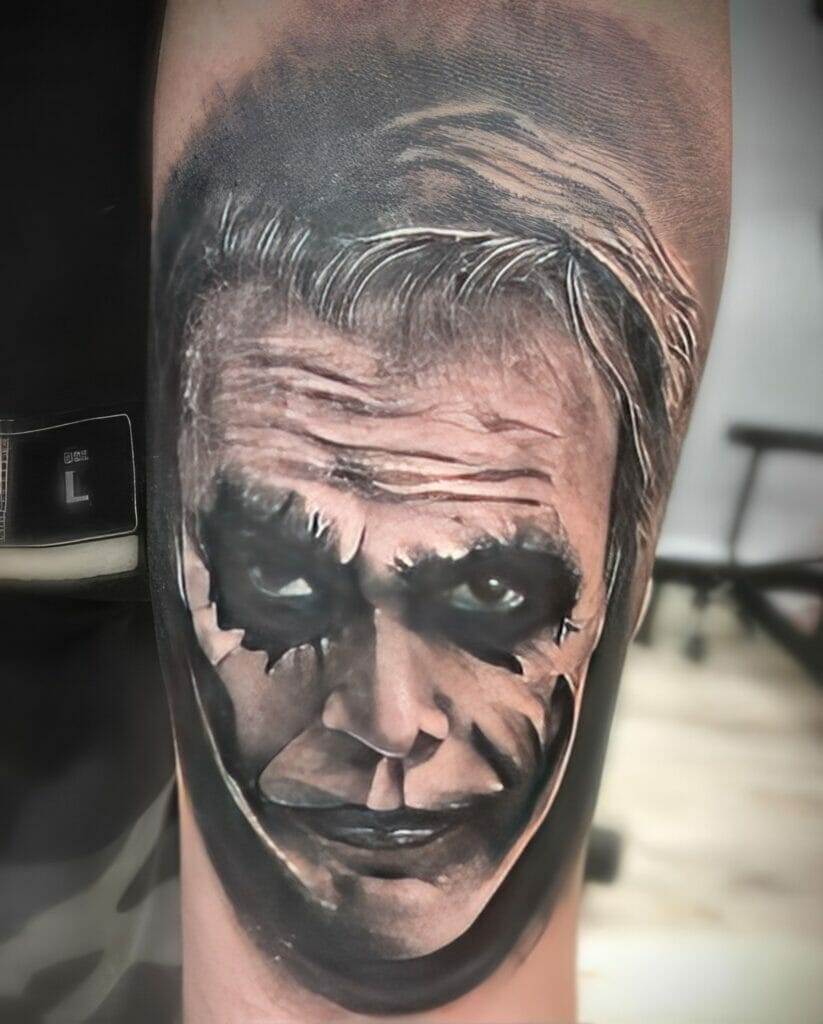 A tattoo of a man's face with heavy makeup resembling a theatrical character, displaying a sinister expression, is inked on the skin.