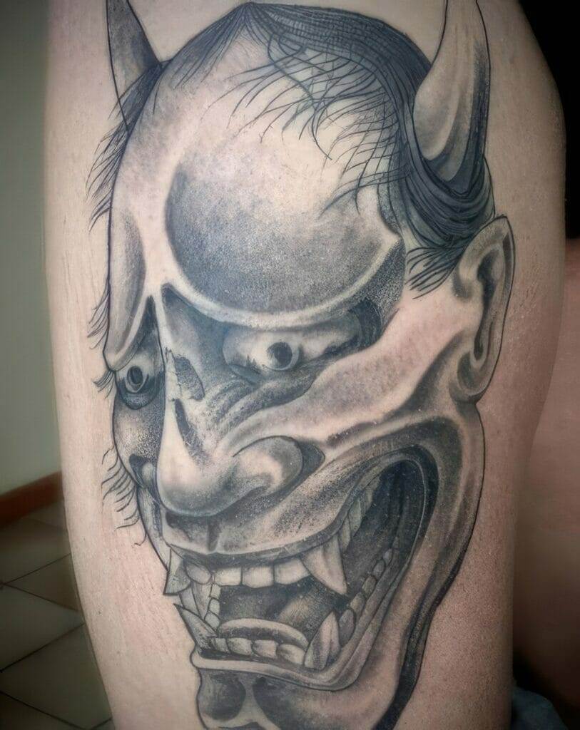 A detailed black and gray tattoo of an Oni mask with sharp teeth and prominent horns is shown on a person's leg.