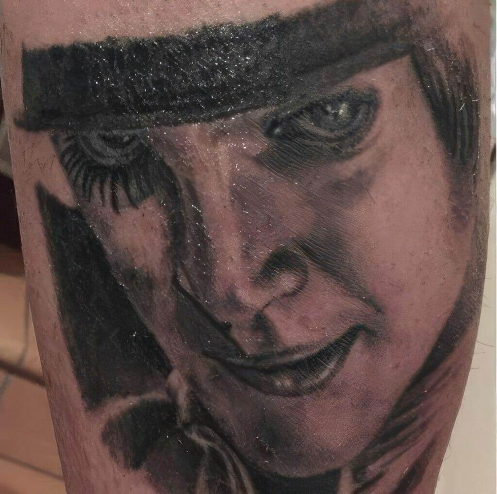 A close-up tattoo on skin depicting a monochromatic portrait of a person wearing dark eye makeup and a hat.