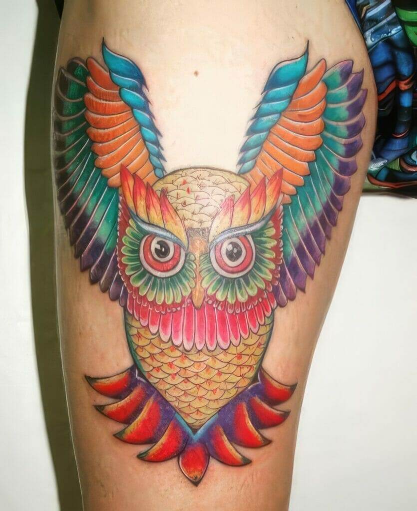 A vibrant, colorful tattoo of an owl with spread wings, featuring intricate details and a blend of red, green, blue, orange, and purple hues on a person's thigh.