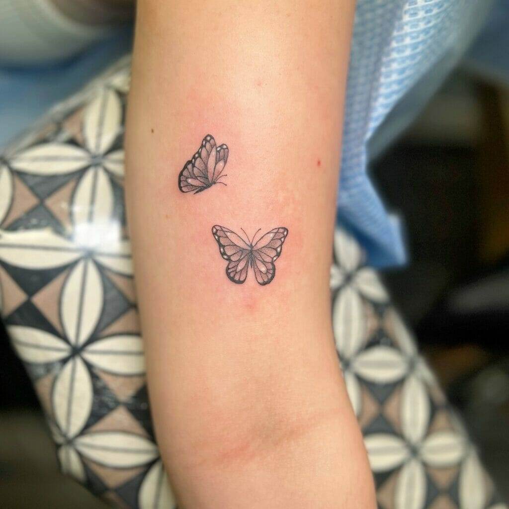 Two black ink butterfly tattoos on a person's arm, with the arm resting on a patterned surface.