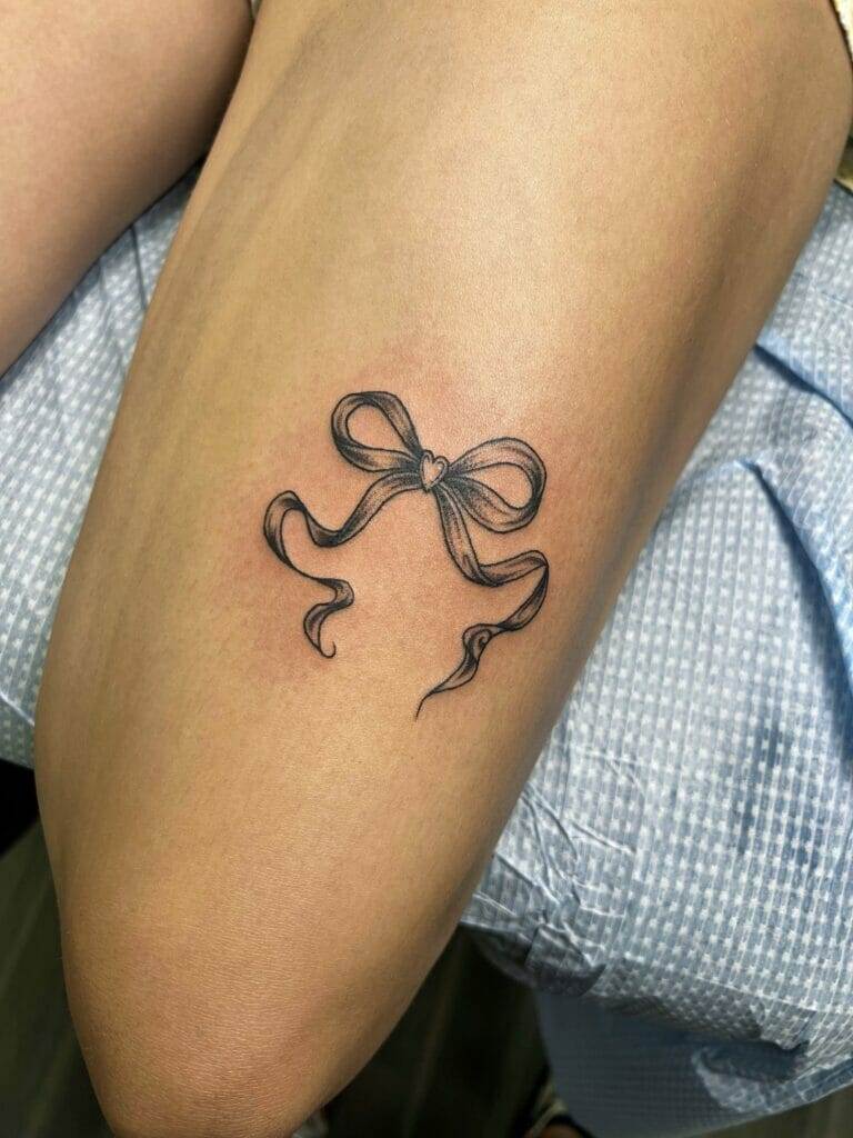 A black and grey tattoo of a ribbon with a small heart in the center is inked on someone's thigh. The surrounding skin appears slightly red, indicating it is freshly done.
