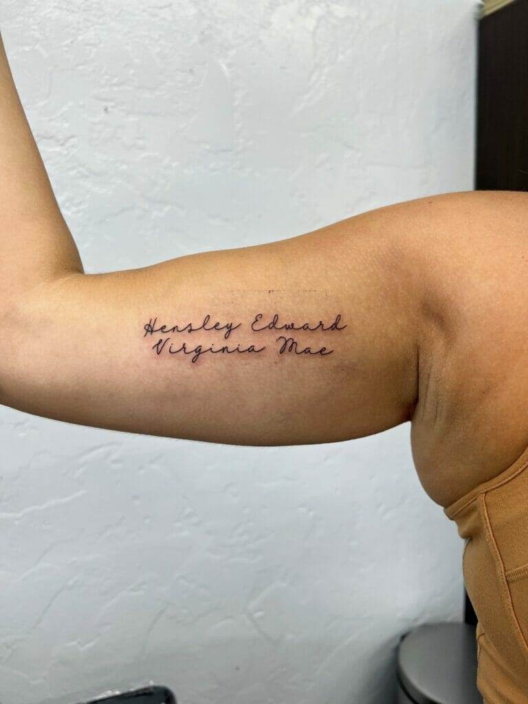 A person's flexed bicep with a tattoo of cursive names "Hensley Edward Virginia Mae" on the inner arm.