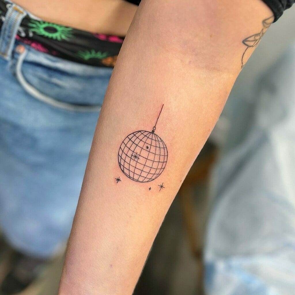 Close-up of an arm showcasing a tattoo of a geometric globe hanging from a string with small dots and stars around it. The background includes a glimpse of blue jeans and patterned clothing.