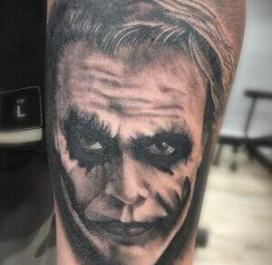 Black and white tattoo of a man's face with heavy makeup, dark eyes, and a sinister smile, resembling a character from a popular comic book.