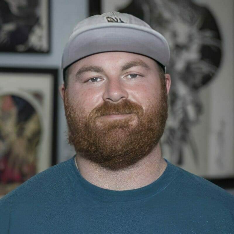 Man with a red beard wearing a gray cap and blue shirt, standing indoors.
