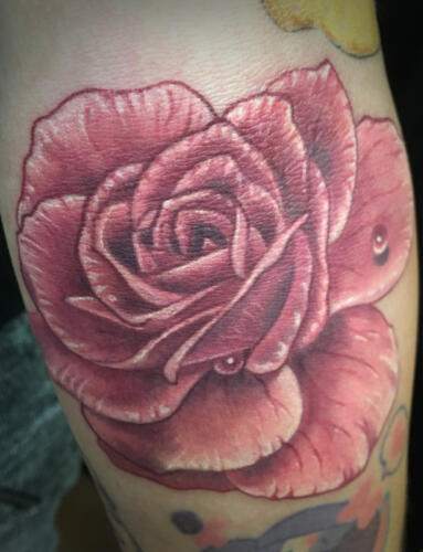 Close-up of a red rose tattoo intricately detailed and shaded, located on a person's skin. The rose is prominently featured and surrounded by smaller, indistinct ink designs.