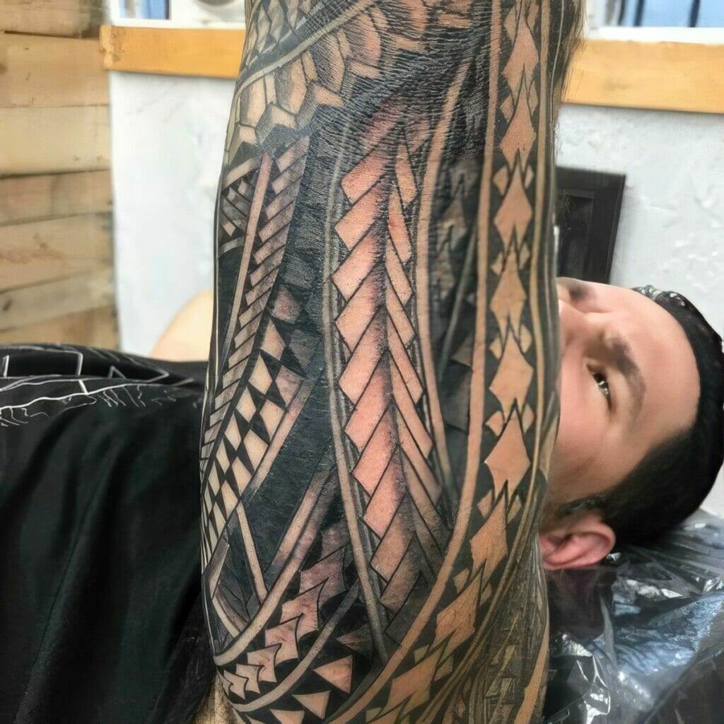 A person reclines while getting a detailed black and gray tattoo featuring geometric and tribal patterns on their arm.