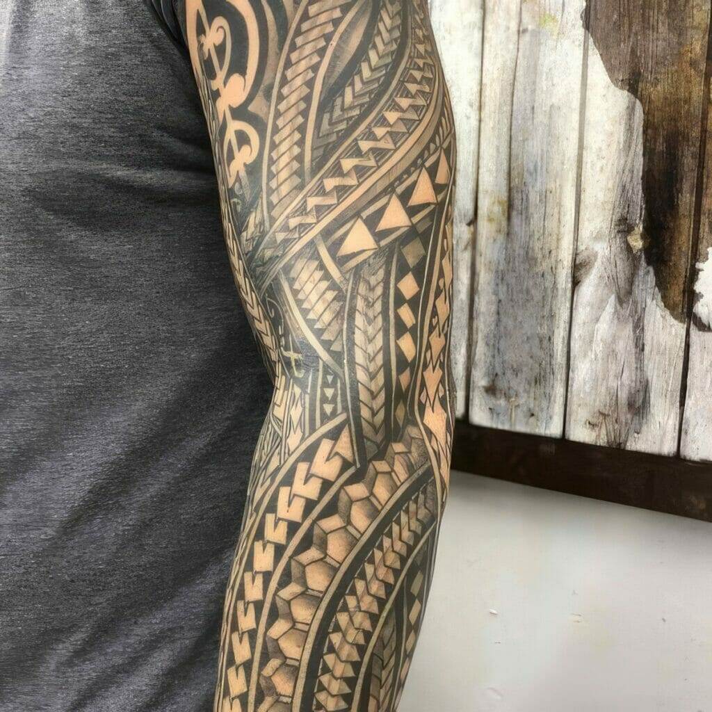 Arm with intricate black tribal tattoo sleeves against a wooden backdrop. The person is wearing a short-sleeved gray shirt.