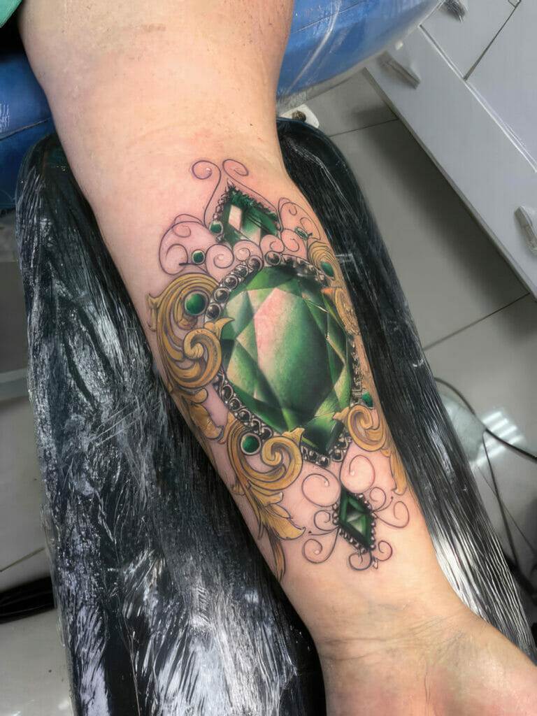 A forearm tattoo featuring a large, detailed green gem surrounded by ornate golden and black decorative elements.