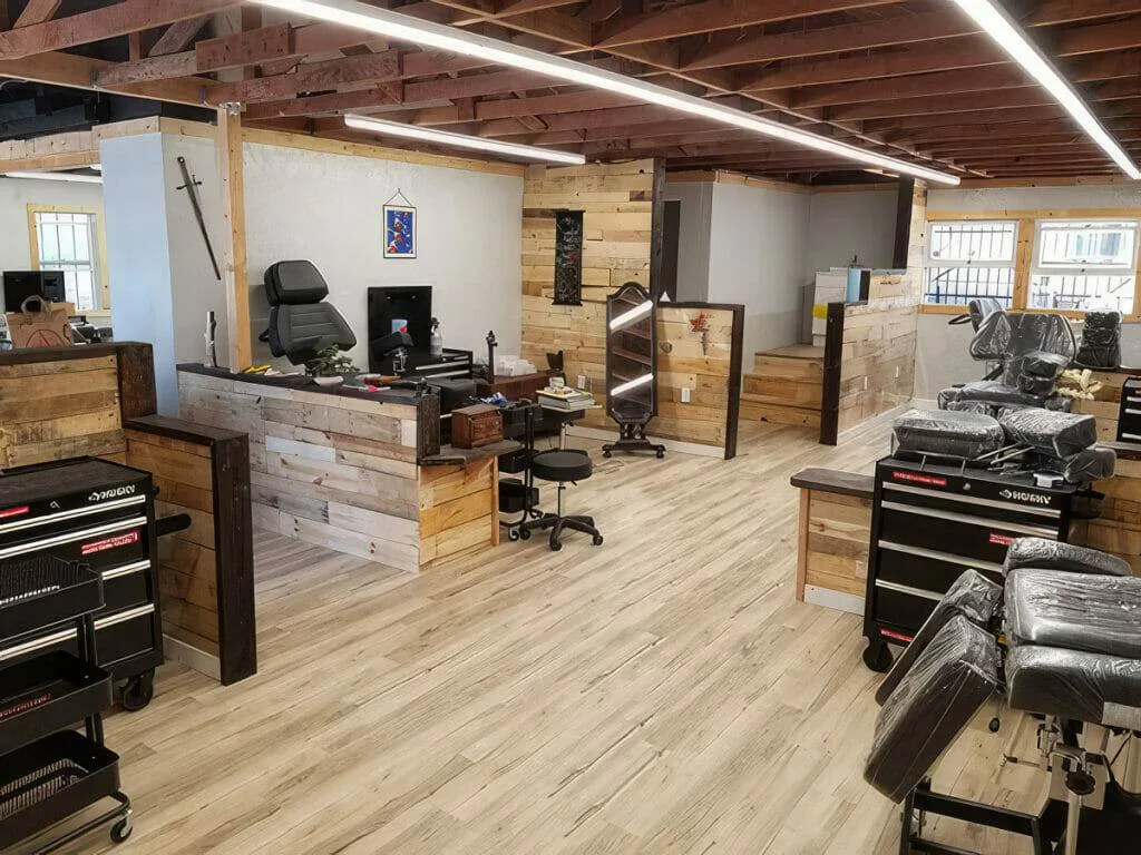 A spacious and modern tattoo parlor with wooden accents, tattoo chairs, workstations, various equipment, and natural light from large windows.