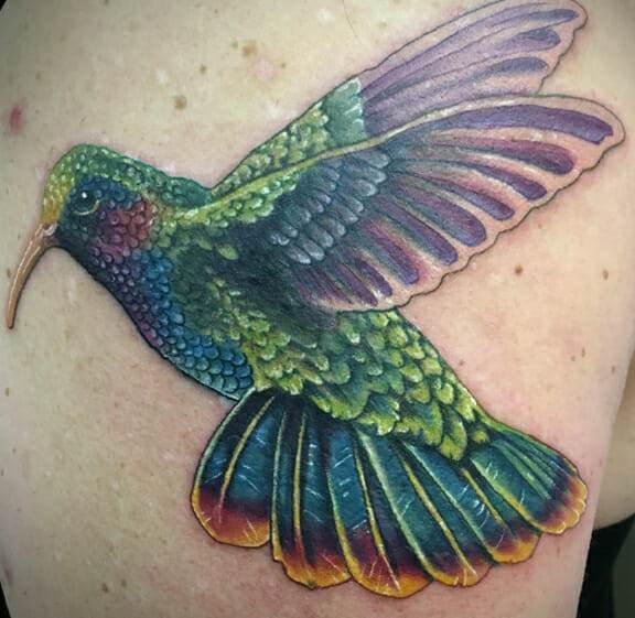 A colorful tattoo of a hummingbird with detailed green, blue, purple, and yellow feathers, seen on a person's upper arm.