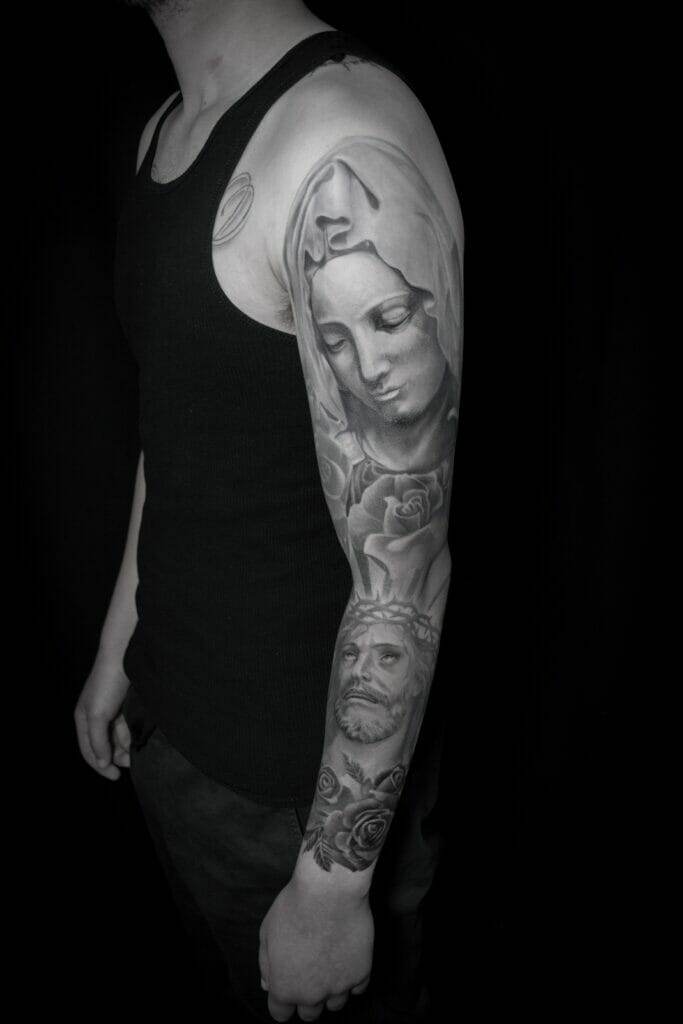 Person in a black tank top showcasing a detailed sleeve tattoo on their left arm, featuring religious imagery including the Virgin Mary and Jesus Christ.