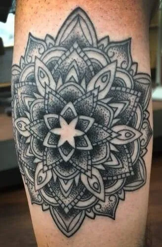 A black and grey geometric mandala tattoo with intricate patterns and shading is shown on light skin.