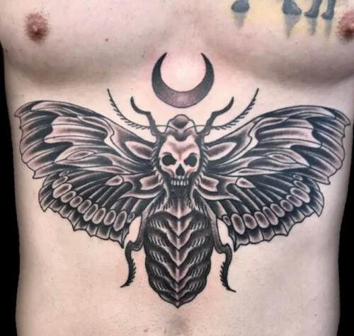 Tattoo of a large moth with skull-like markings on its thorax and an upward-facing crescent moon above it, located on a person's chest.