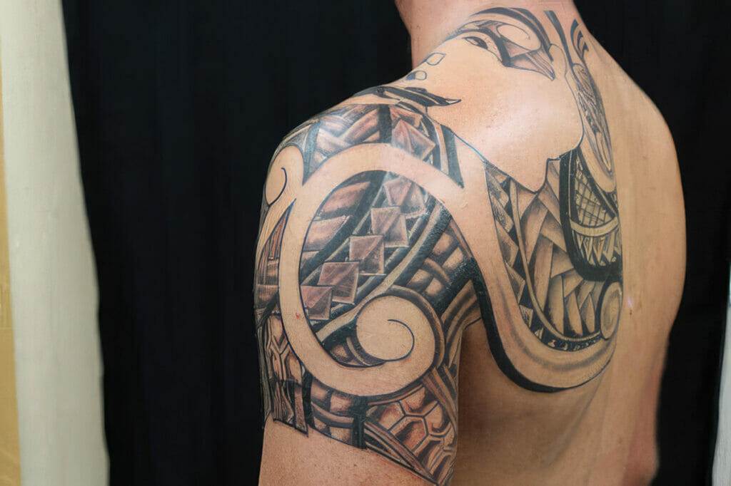 A person has a large, intricate tattoo covering their shoulder and upper arm, featuring geometric and abstract designs with black and skin-colored inks.