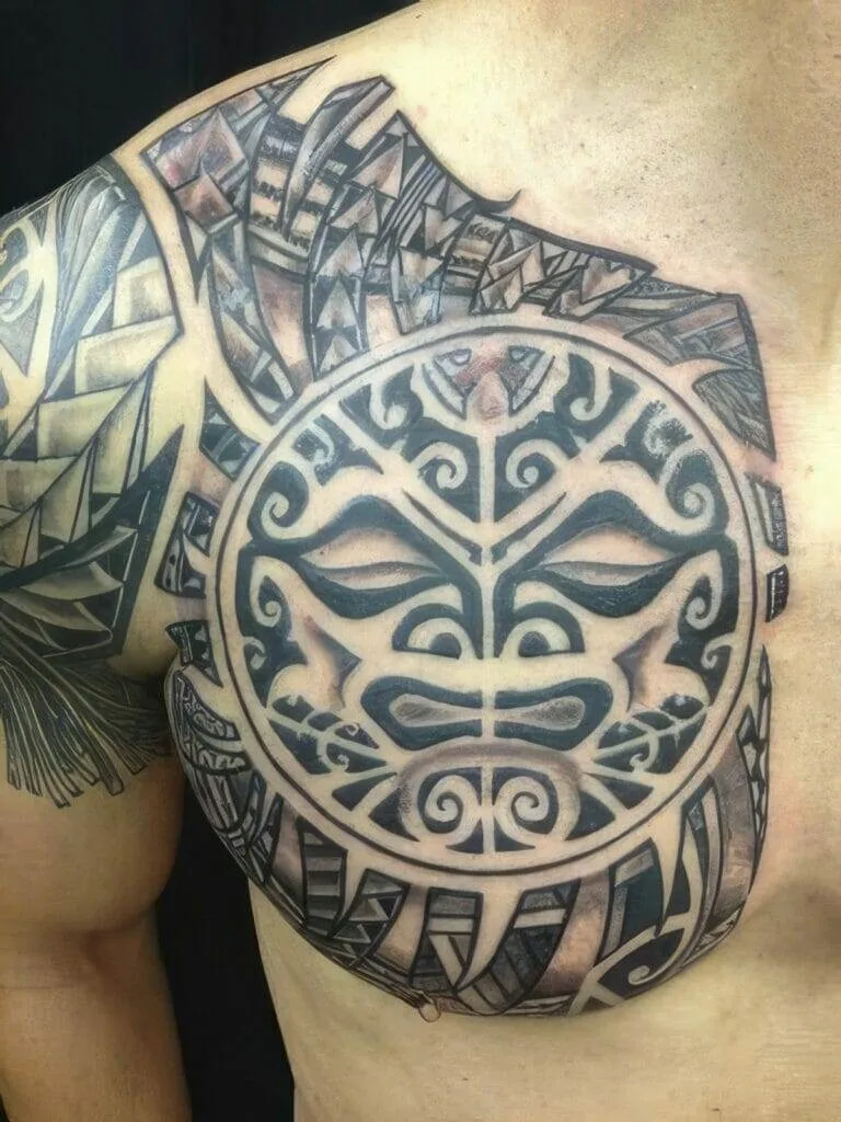 A close-up of a tribal tattoo covering part of a person's chest and shoulder. The design features a central circular pattern with stylized faces and geometric patterns extending outward.
