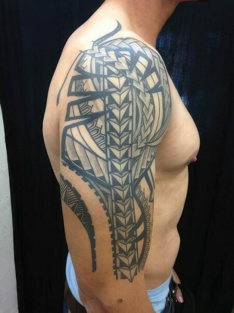 A person with an intricate geometric tattoo covering their shoulder and upper arm, standing against a dark background.