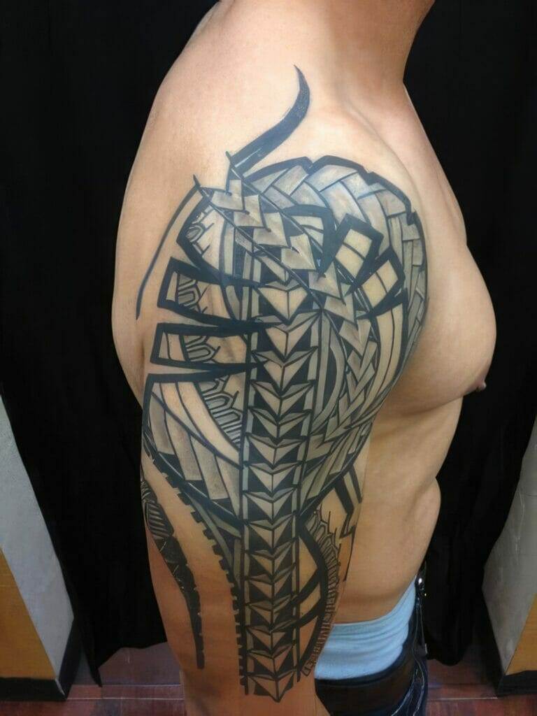 A person with a large, intricate tribal tattoo covering their upper arm and shoulder, featuring bold geometric and angular designs in black ink. The person stands against a dark background.