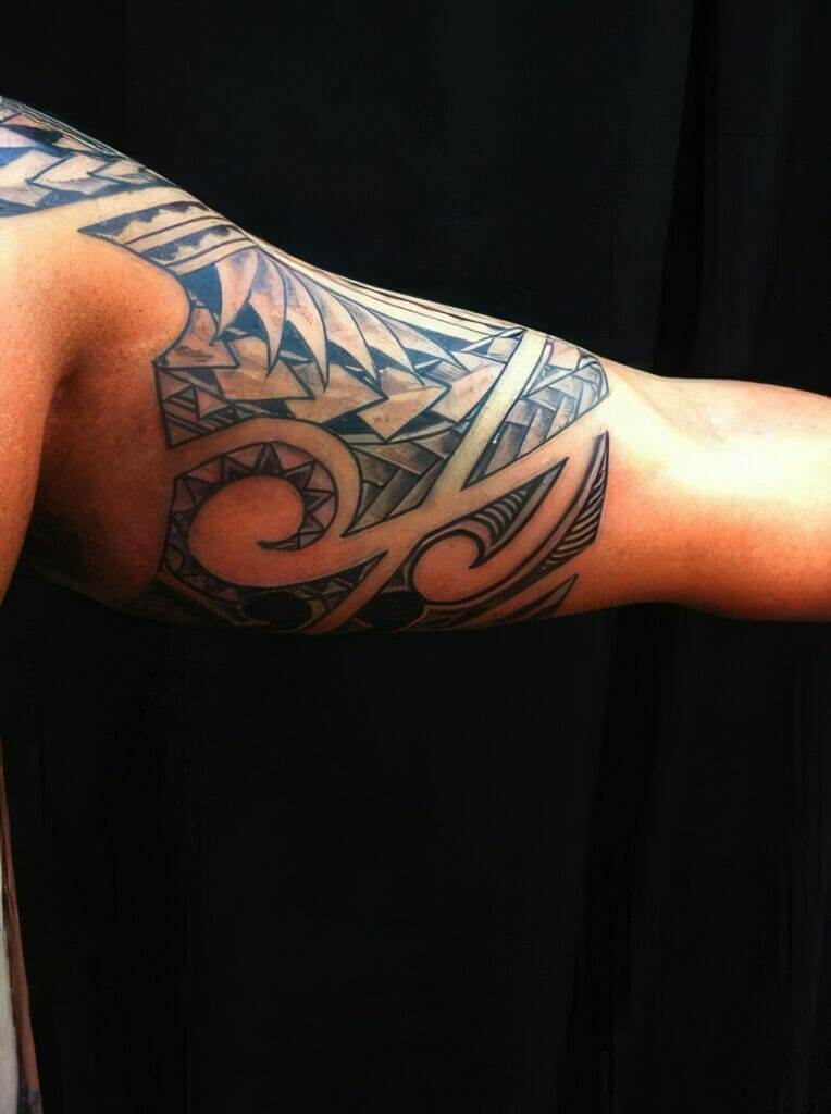 A close-up of a person's upper arm displaying a detailed black tribal tattoo against a black background.