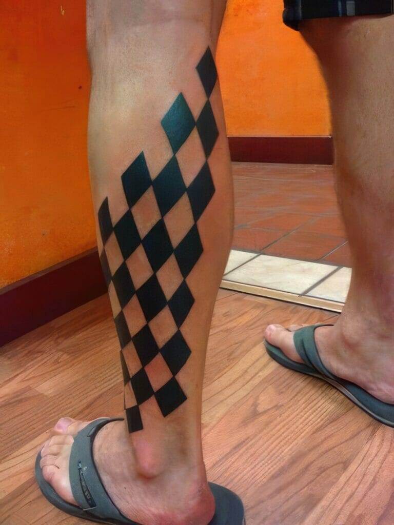 A person's lower leg with a large black and dark orange checkered tattoo design. The person is wearing flip-flops and standing indoors on a wooden floor.