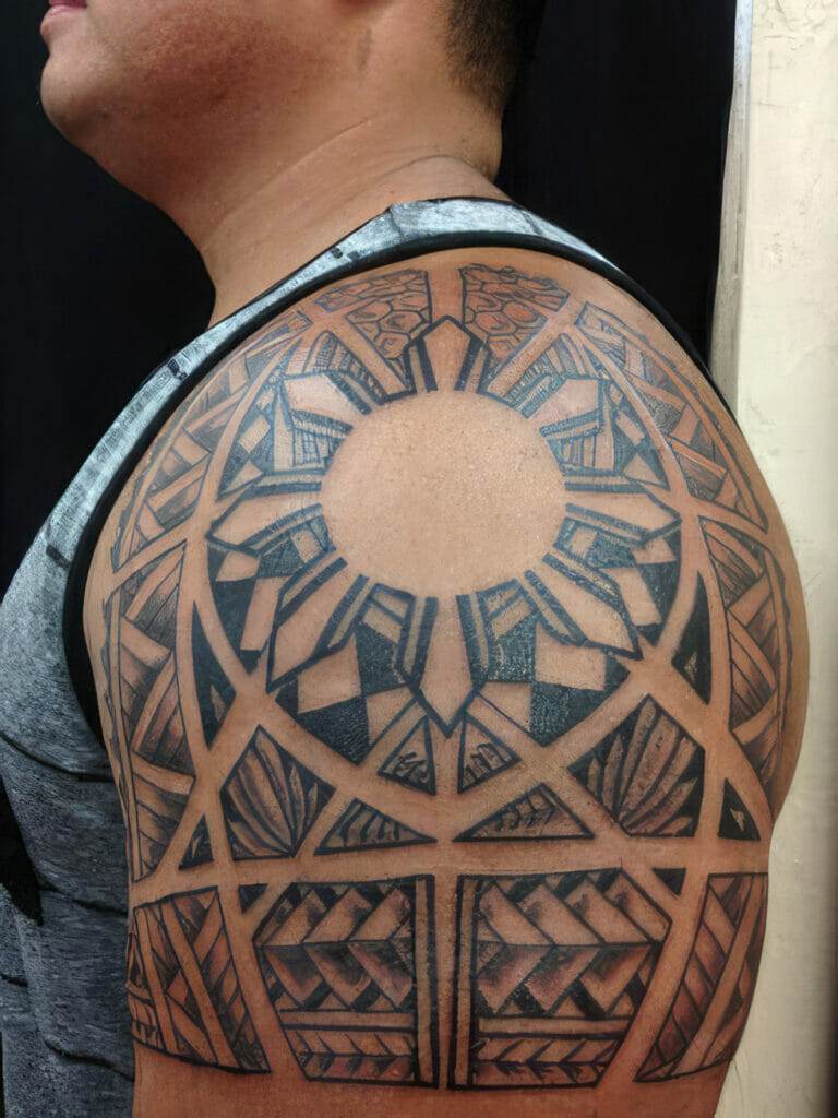 A person with an intricate geometric tattoo covering their shoulder and upper arm. The tattoo features a circular design at the center and various triangular and angular patterns.