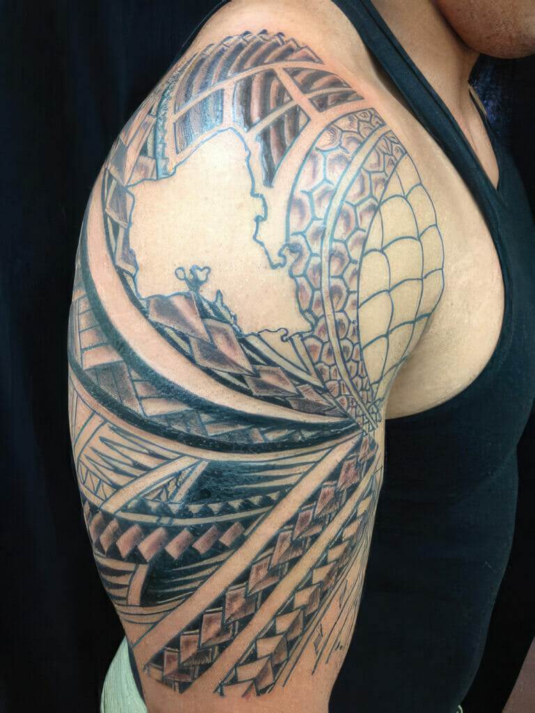 A person's upper arm with intricate black ink geometric and tribal tattoo patterns, including elements such as triangles, lines, and curved shapes.