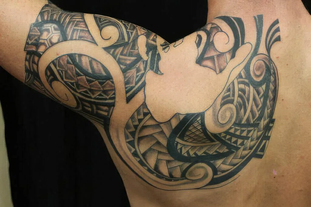 Lower arm and upper back displaying an intricate, tribal-patterned tattoo, primarily in black ink, with various geometric shapes and swirls.