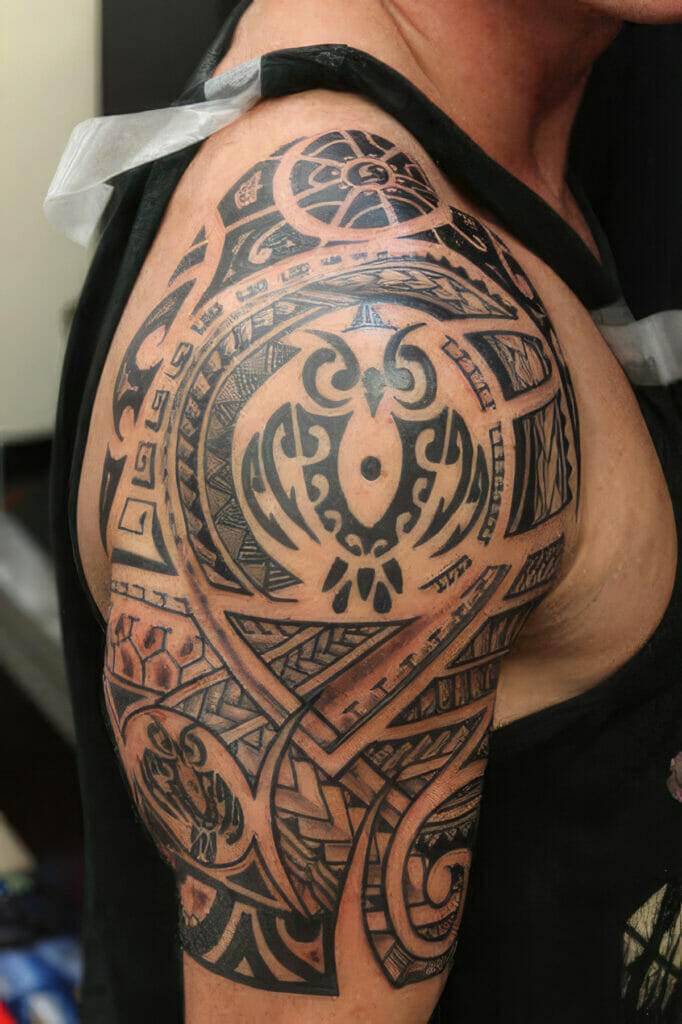 A person with an intricate black tribal tattoo covering their upper arm and shoulder. The tattoo design features geometric patterns and symmetrical shapes.